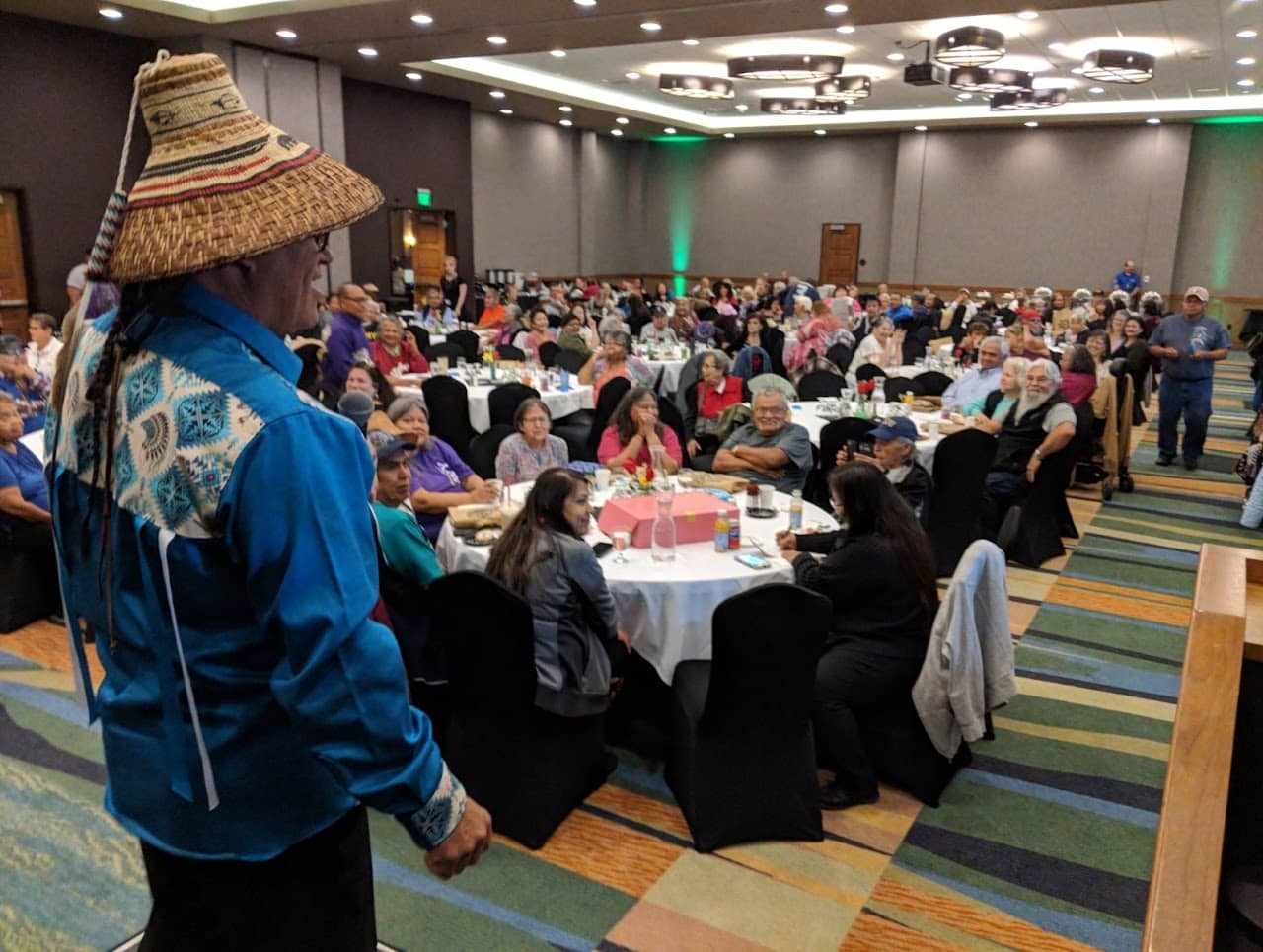 unnamed-2 A man whearing a blue long sleeve dress shirt and cedar hat stands in front of a conference room filled with round tables and people eating lunch.